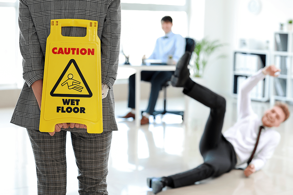 Man slipping on the floor in an office with a coworker holding a wet floor sign behind their back | Hiring A Slip And Fall Lawyer